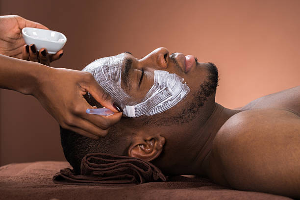Therapist Applying Face Mask To Young African Man In Spa